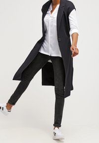 Lång svart väst med öppen front, bärs över en vit skjorta med knappar och svarta jeans. Modellen har på sig vita sneakers med svarta detaljer.