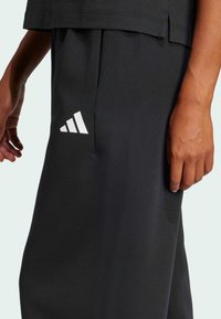 Juodi sportiniai kelnių iš minkštos medžiagos, su balta Adidas logo ant kairio šlaunies, laisvo silueto ir šoninėmis kišenėmis.