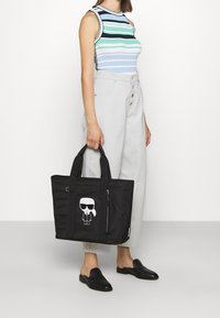 Zwarte schoudertas van duurzame stof, met een cartoonafbeelding, een ritsvak en dubbele handvatten. Stevig, gestructureerd ontwerp.