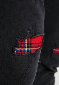 Denim negro con parches desgastados que revelan una tela de cuadros roja, amarilla y azul debajo en la zona del muslo de los pantalones.