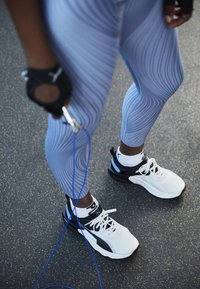 Lichtblauwe legging met golvende patronen; zwart-witte sportschoenen met blauwe accenten; springtouw in de hand, staande op een textuuroppervlak.