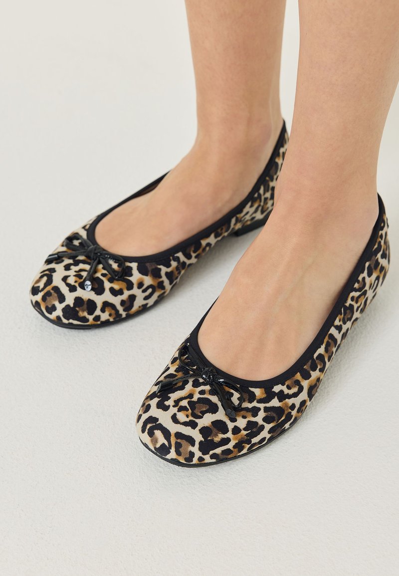 Pieds portant des ballerines imprimé léopard avec une bordure noire et de petits nœuds décoratifs sur un fond de sol clair.