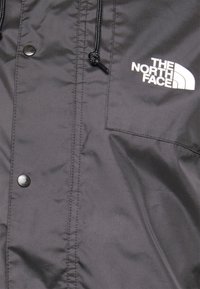 Tissu de veste noire avec des boutons-pression, un cordon de serrage et un logo blanc "The North Face" sur la partie supérieure droite de la poitrine.
