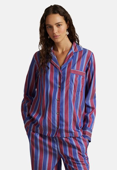 Femme portant un ensemble de pyjama à boutons rayé verticalement bleu et bordeaux avec col et poche avant, debout les mains dans les poches du pantalon.
