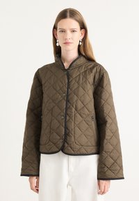 Veste matelassée marron avec un col arrondi, des bordures noires, des boutons-pression et des poches latérales. Le matériau semble rembourré avec une texture lisse.