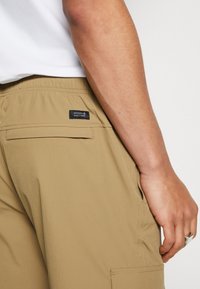 Shorts cargo beiges avec une poche arrière ornée d'une étiquette de marque noire. Tissu lisse avec une coupe décontractée et une taille élastique.