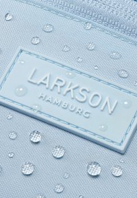 Vaaleansininen kangaskassi, jossa on teksturoitu pinta ja useita vesipisaroita. Kassin reunassa näkyy kohotettu valkoinen "LARKSON HAMBURG" -logo.
