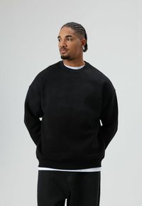 Zwarte oversized sweatshirt met een ronde hals, voorzien van textuurelementen en een losse pasvorm, gecombineerd met donkergrijze jeans en een wit T-shirt eronder.