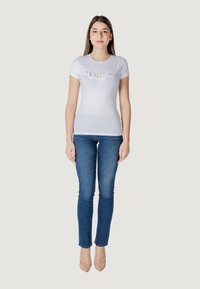 Wit aansluitend t-shirt met "Guess" logo in zilveren glitter, gecombineerd met donkerblauwe skinny jeans en nude hoge hakken, staande op een effen achtergrond.