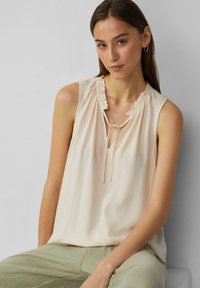 Helle beige ärmellose Bluse mit gerafftem Ausschnitt und Schlüsselloch-Bindedetail, aus glattem Stoff gefertigt, kombiniert mit strukturierten grünen Hosen.