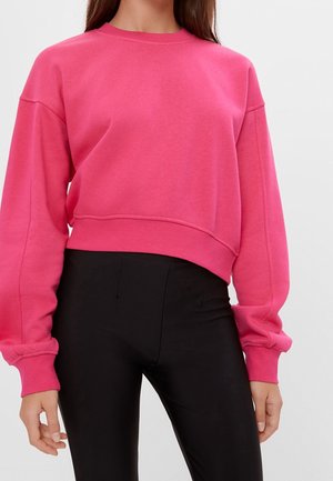 Nainen, jolla on päällään kirkkaan pinkki pitkähihainen lyhyt toppi ja mustat korkeavyötäröiset legginsit, yksinkertaisen taustan edessä.