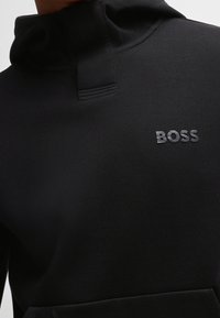 Felpa nera realizzata in tessuto testurizzato. Presenta un colletto alto, tasca frontale e la parola "BOSS" in lettere grigie sul petto.