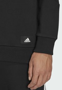 Felpa nera con polsini a coste. Presenta un piccolo logo Adidas bianco su una patch in tessuto nero. Texture morbida e design rilassato.