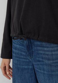 Zwarte top met lange mouwen, voorzien van een gerimpelde, verstelbare zoom en een subtiele textuur, gecombineerd met blauwe spijkerbroek met een vervaagd patroon.