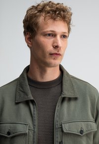 Olijfgroene jas met twee voorzakken, met een kraag met knopen, gedragen over een gestructureerd donkerbruin sweater met een ronde hals.