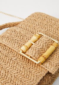 Borsa a tracolla intrecciata in paglia beige con superficie texture. Dotata di una chiusura in bambù e hardware color oro. Inclusa tracolla a catena.
