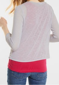 Kvinna i ett genomskinligt ljusgrått långärmat topp över en röd tröja och blå jeans, vänd bort med vänster arm böjd.