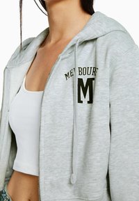 Felpa grigia con zip e vestibilità rilassata, caratterizzata da un logo che recita "MELBOURNE" e una "M" maiuscola in stampa scura. Materiale misto cotone.