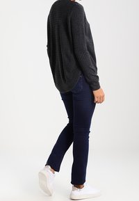 Person trägt einen dunkelgrauen, gerippten Pullover, dunkelblaue Jeans und weiße Sneakers und steht mit einem leicht angehobenen Fuß vor weißem Hintergrund.