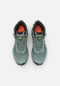 Scarpe atletiche in mesh verde e sintetico con sovrapposizioni testurizzate, accenti riflettenti e soletti arancioni, caratterizzate da una suola nera robusta.