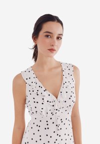 Witte jurk met zwarte stippen, voorzien van een ruchesinspectie en een strikdetail aan de voorkant, gemaakt van lichte stof.