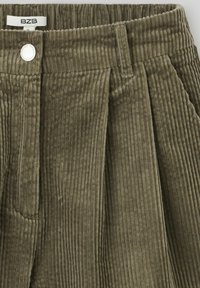 Pantalons en velours côtelé vert olive avec des plis verticaux, dotés d'une fermeture à bouton et d'un tissu texturé à côtes. Étiquette de marque visible à la taille.