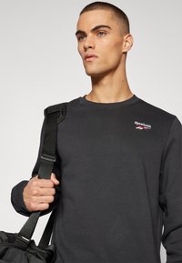 Sweat-shirt noir avec un col rond, logo Reebok sur la poitrine et tissu texturé. Le modèle tient un sac avec une bandoulière sur son épaule.