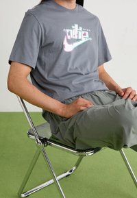 Šedé bavlněné tričko s barevným designem loga Nike, spojené se světle zelenými tepláky, sedící na průhledné židli na zelené podlaze.