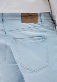 Lyseblå denimjeans med en tekstureret overflade, synlige sømme og et brunt lædermærke på den bagerste talje.