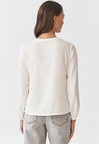 Lys beige bluse med lange ærmer, glat tekstur, rund halsudskæring og samlede manchetter, parret med lysegrå, falmede denim jeans.
