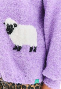 Fioletowy miękki sweter z białą i czarną owcą na drutach, noszony z legginsami w panterkę, ukazujący szczegóły rękawów i dołu.