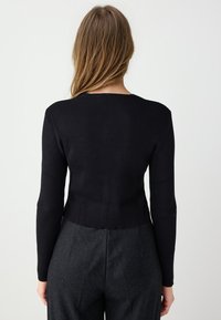 Haut noir à manches longues côtelé avec un col rond, présentant un design court et un matériau texturé. Porté avec un pantalon gris foncé.