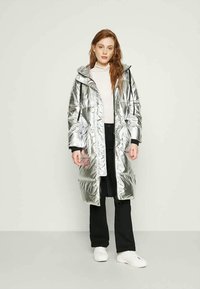 Casaco puffer prateado com capuz, fecho de correr e cordão. Apresenta um padrão acolchoado e bolsos laterais. Usado sobre uma blusa de cor clara.