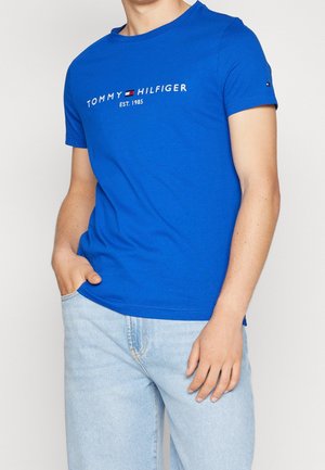 Mężczyzna ubrany w jaskrawo niebieską koszulkę Tommy Hilfiger z logo oraz jasnoniebieskie dżinsy, jedna ręka w kieszeni, na jednolitym tle.