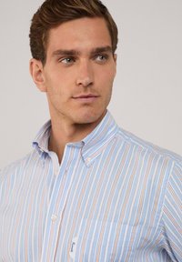 Camisa de color azul claro con cuello y rayas verticales en naranja y blanco. Cuenta con un bolsillo en el pecho y cuello abotonado. Tejido de mezcla de algodón.