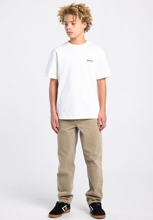 Adolescent aux cheveux bouclés portant un t-shirt blanc "Chicago", un pantalon beige et des baskets noires, debout devant un fond clair uni.