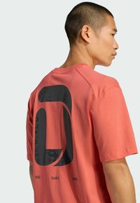 Camiseta coral de manga corta con un gran gráfico negro en la parte trasera con el texto "2026 TEAM D ADIDAS", hecha de material de algodón ligero.