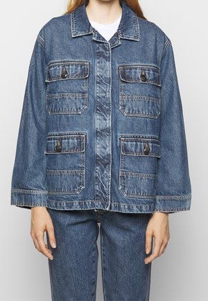 Person iført en blå denimjakke med fire knaplåse lommer og matchende denim jeans, hænderne afslappede ved siderne.