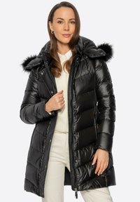 Mujer con un abrigo largo negro acolchado con capucha con borde de piel, suéter crema y pantalones crema, de pie frente a un fondo blanco.