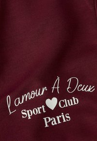 Tela burdeos con una textura suave que presenta texto impreso en blanco: "L'amour À Deux Sport Club Paris" y un icono de corazón.