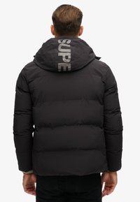 Schwarze Daunenjacke mit Kapuze, die ein großes "SUPER"-Logo auf dem Rücken trägt. Das Design ist gesteppt und hat eine glatte Textur.