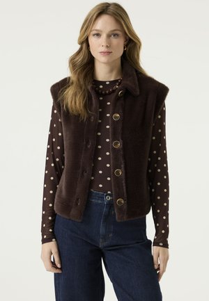 Femme portant un gilet sans manches en fausse fourrure marron foncé sur un top marron à pois à manches longues et un jean bleu, debout devant un fond blanc.
