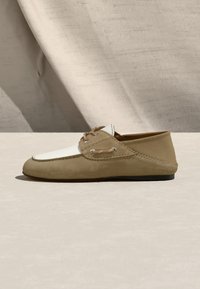 Mocasín de ante beige y blanco con detalle de cordones y ojales metálicos, exhibido sobre una superficie neutra con un fondo de tela suave.