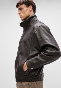 Pruun nahkjope kõrge kraega, küljetaskudega ja kogutud allääriga. Silmapaistev tekstuur nähtavate õmbluste detailidega.