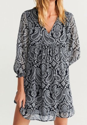 Frau trägt ein langärmeliges Kleid mit V-Ausschnitt und geraffter Taille in einem schwarz-weißen Paisley-Muster.