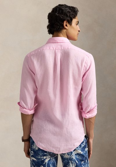 Férfi háttal, világos rózsaszín hosszú ujjú ingben, felhajtott mandzsettákkal és kék, trópusi mintás rövidnadrágban.