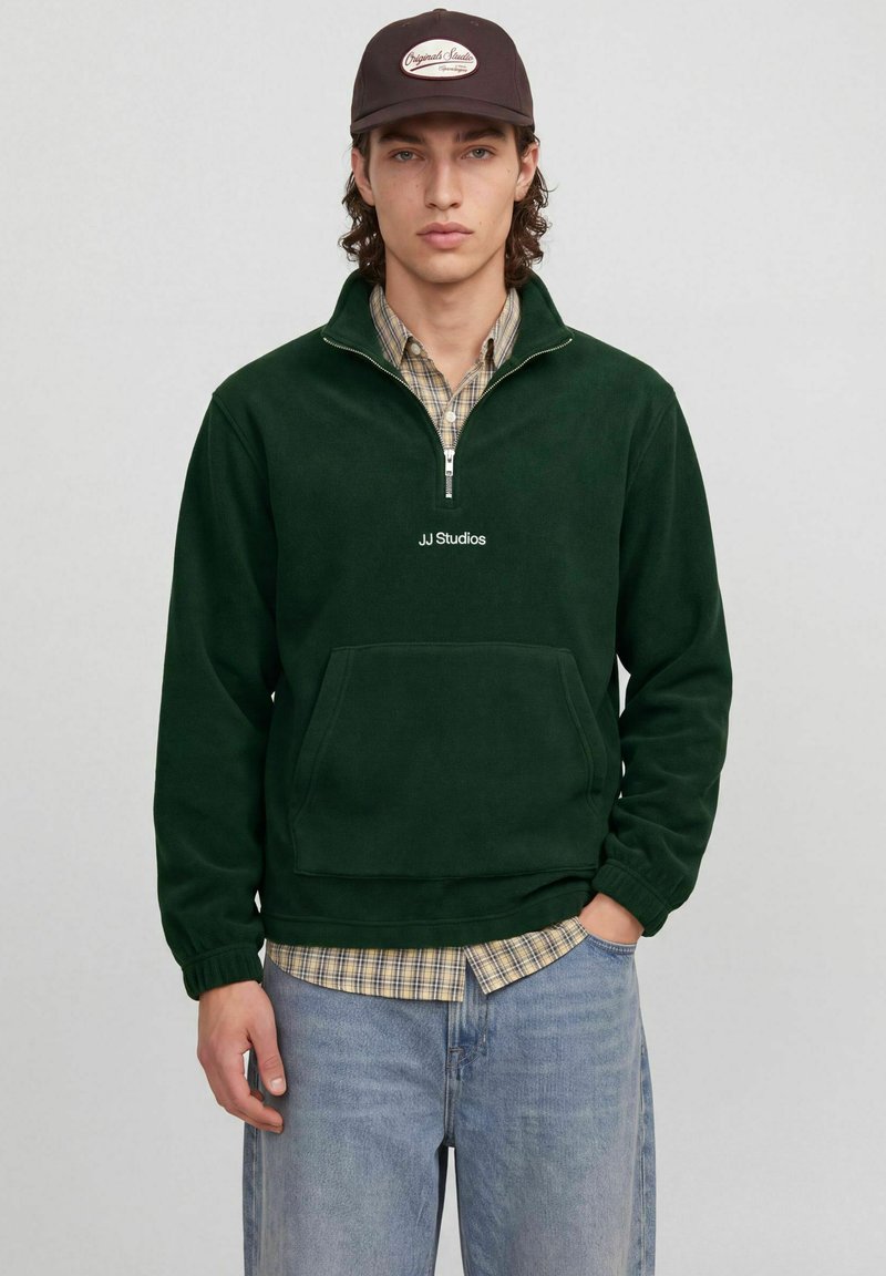 Jeune homme portant une polaire quarter-zip JJ Studios vert foncé, une chemise à carreaux, un jean bleu et une casquette marron foncé avec un logo ovale.