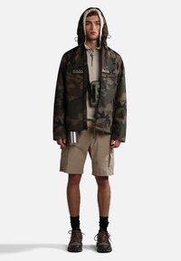 Giacca mimetica con cappuccio, felpa beige, pantaloni corti beige e scarpe da trekking nere. Include una borraccia argentata e una borsa a tracolla.