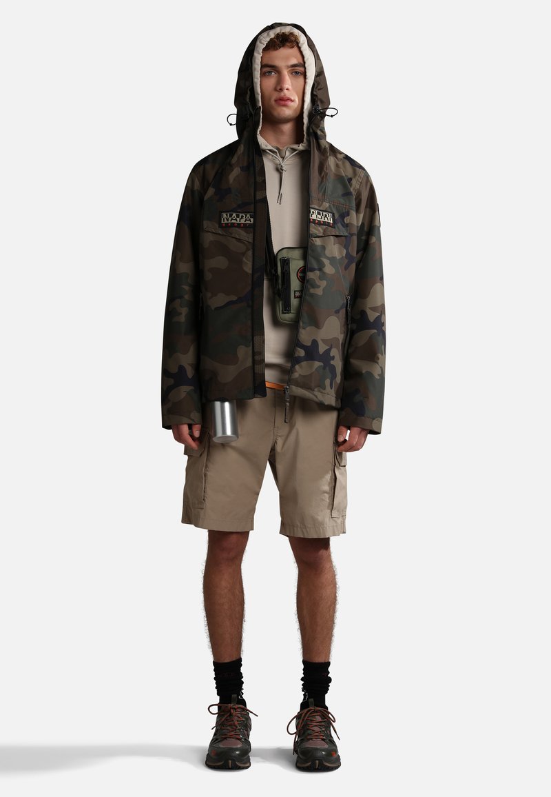 Tarnjacke mit Kapuze, beiges Sweatshirt, beiger Shorts und schwarzen Wanderschuhen. Enthält eine silberne Wasserflasche und eine Umhängetasche.