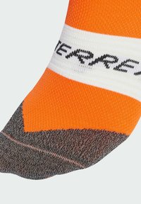 Oranžová ponožka s bielym pruhom s čiernym textom. Špička je tmavo šedá s textúrovaným povrchom. Dizajn je športový a priliehavý.